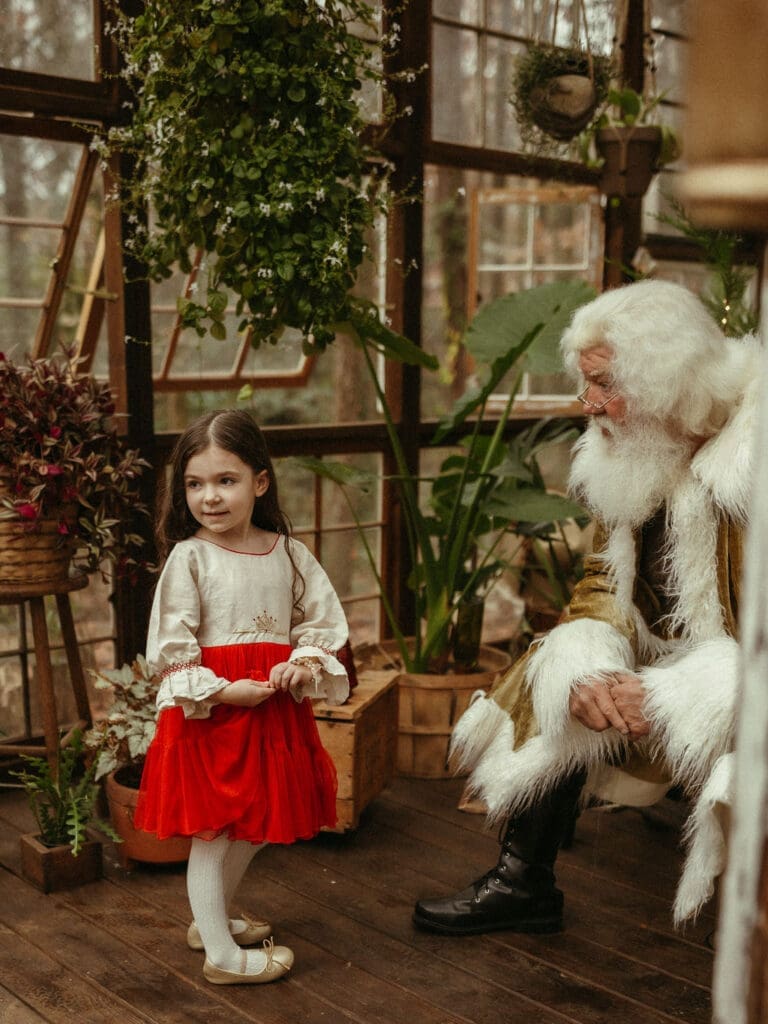 A toddler girl in a bright red skirt standing with groovy Santa and looking back over her shoulder. 