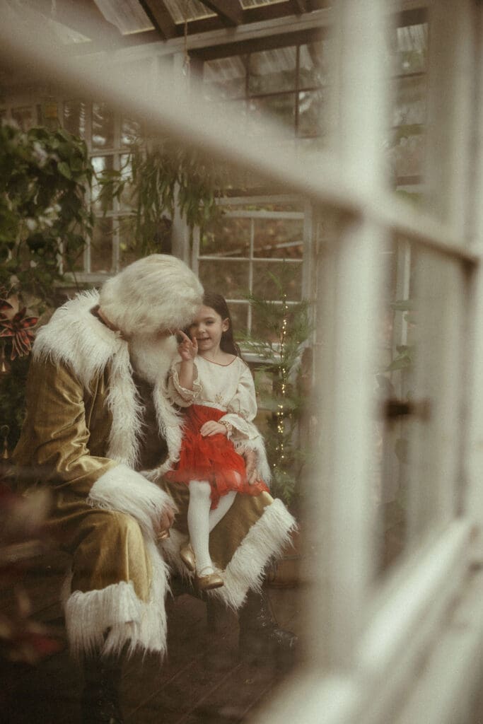 A little girl in a bright red skirt sitting in Santa's lap and poking his nose. 