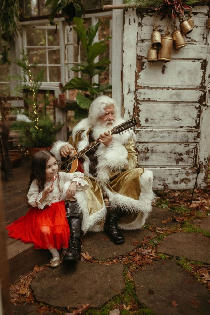 A little girl in a bright red skirt leaning on Santa's leg while he strums the guitar at Das Hinterhaus Studio. 