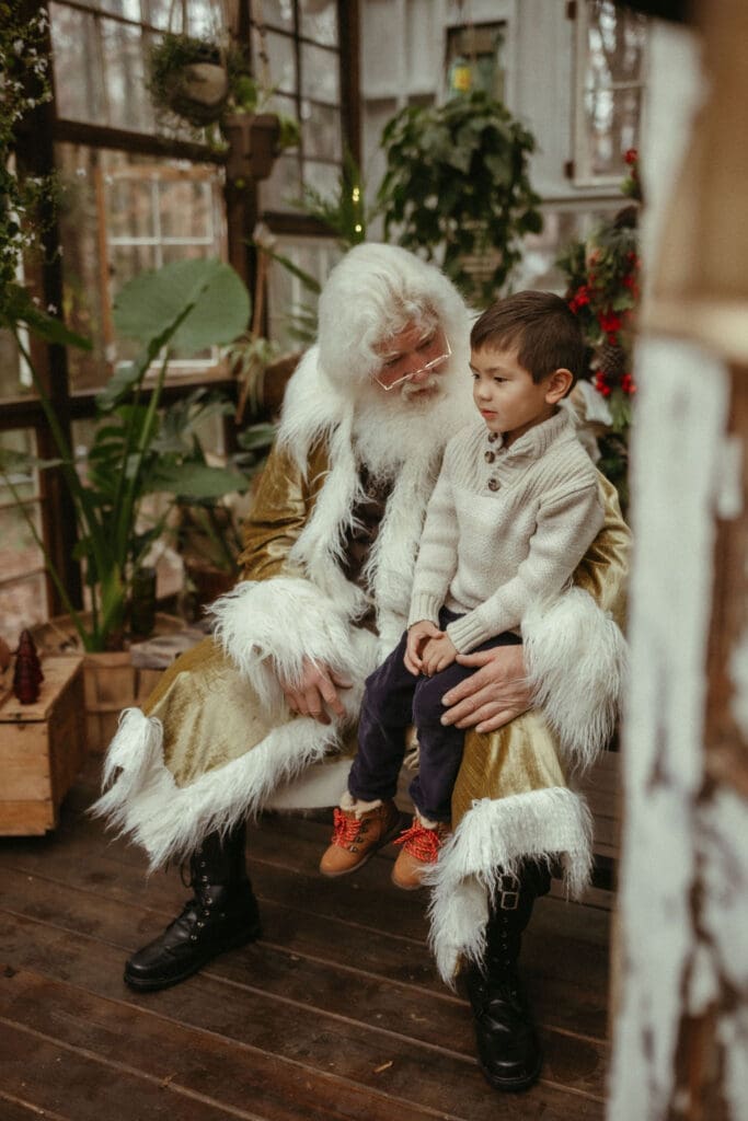 A young boy in a white sweater sitting in The Singing Santa of Atlanta's lap at Das Hinterhaus Studio. 