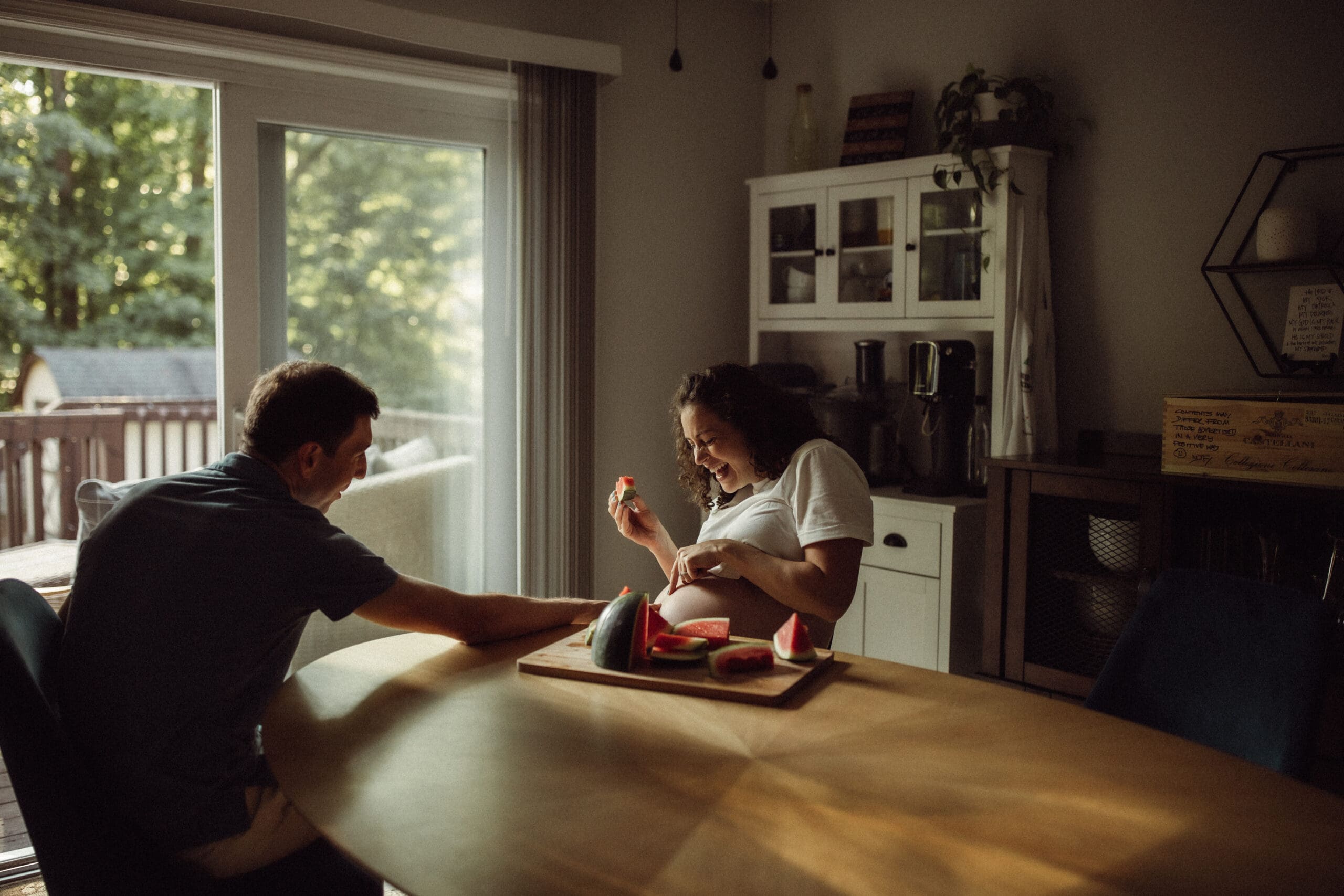 An expecting couple sitting at their dining room table laughing and eating watermelon.