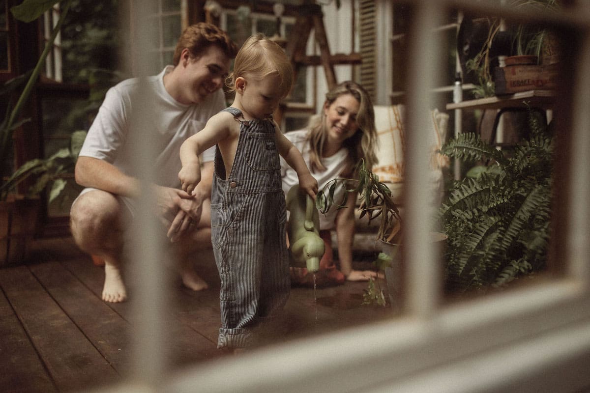 A toddler boy wearing pint striped overalls plays with a watering can as his parents watch at Das Hinterhaus in Marietta, GA.