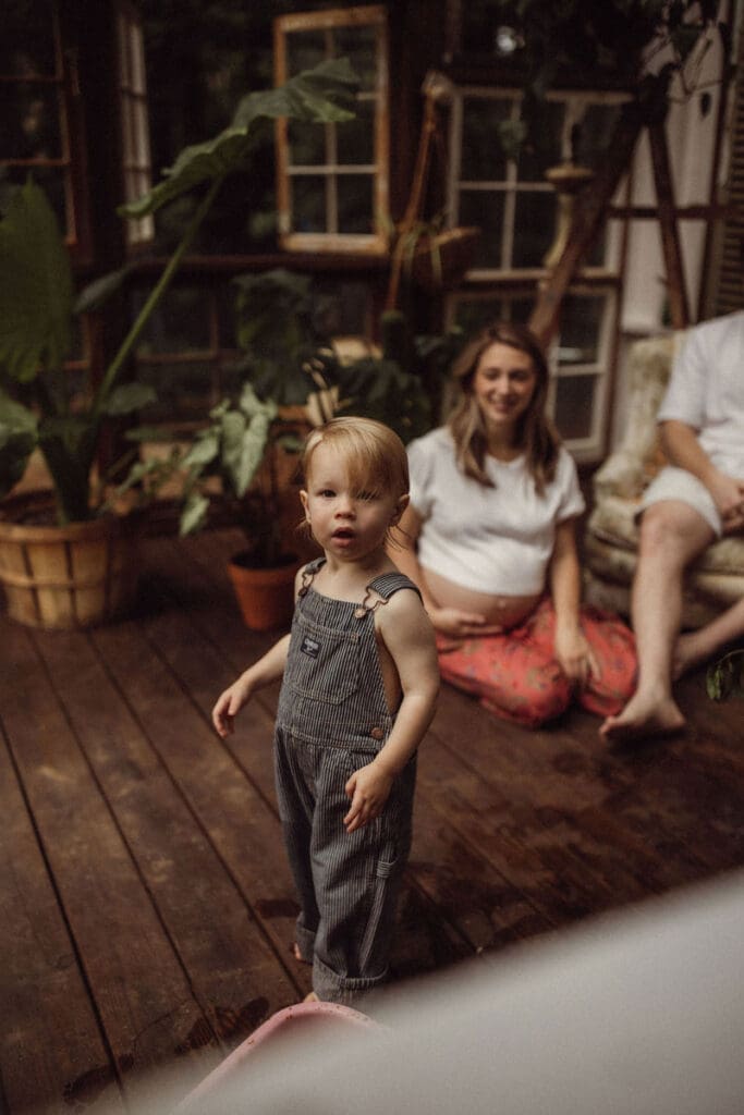 A little blonde toddler boy wearing overalls looking to the camera as his parents watch in the background. 