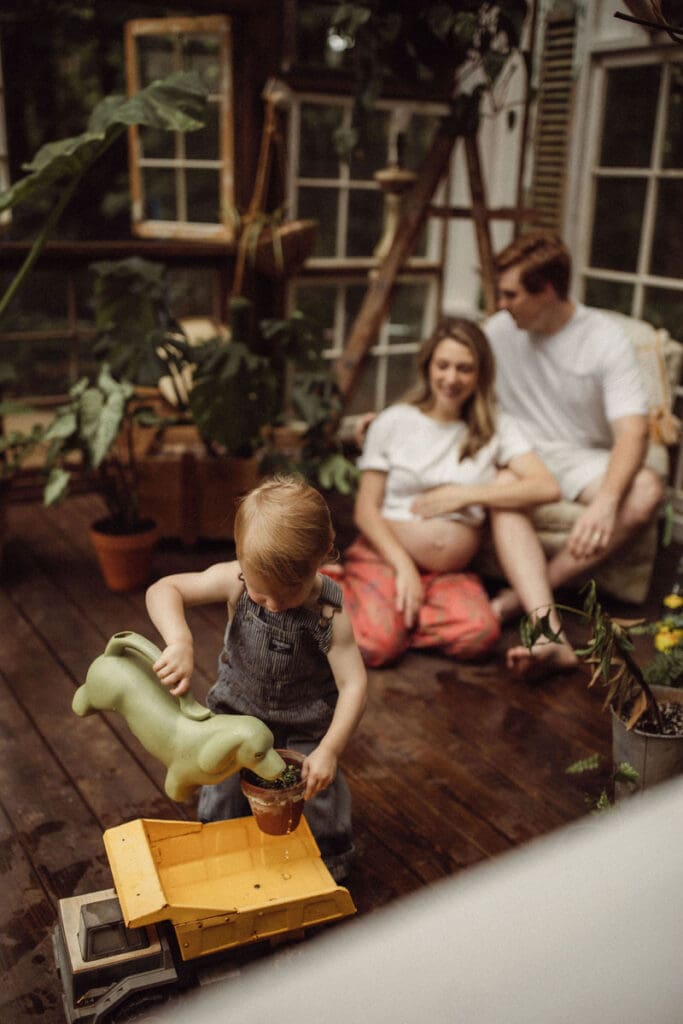 An expecting couple sitting and watching their toddler boy water plants at Das Hinterhaus. 