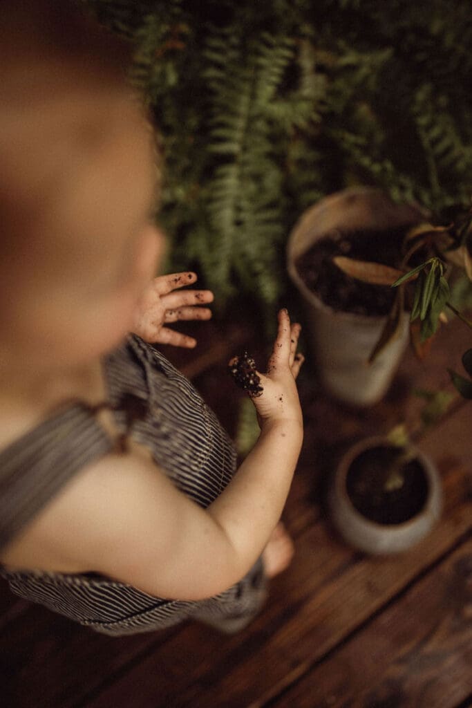 A blonde toddler looking down at their soil covered fingers. 