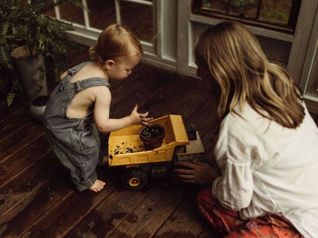 A toddler boy playing in the soil of a potted plant in a dump truck toy while his mom plays along. 