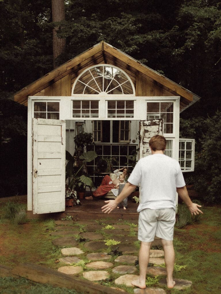 A father holding out his hands and walking to his pregnant wife and toddler who are sitting in a chair at Das Hinterhouse studio. 