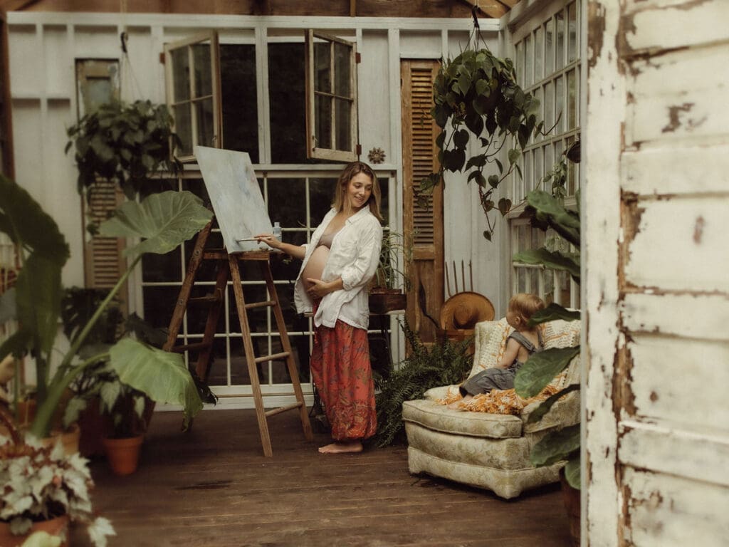 A pregnant mom holding her belly and painting on an easel while looking back at her toddler who is watching. 