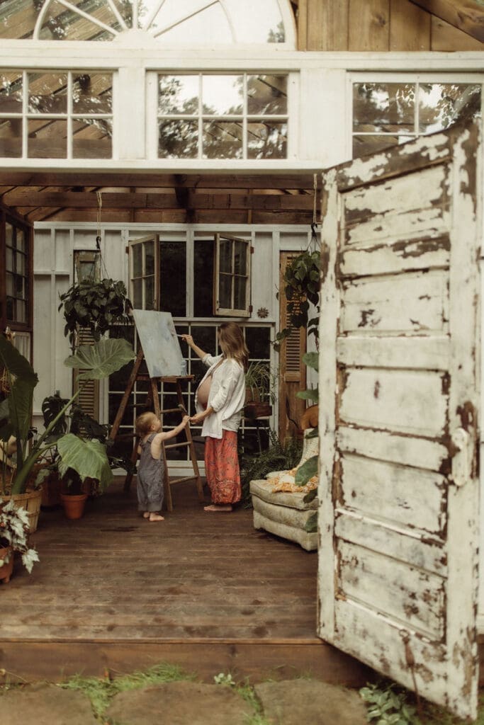 A pregnant mom holding her belly and painting on an easel while holding hands with her toddler. 