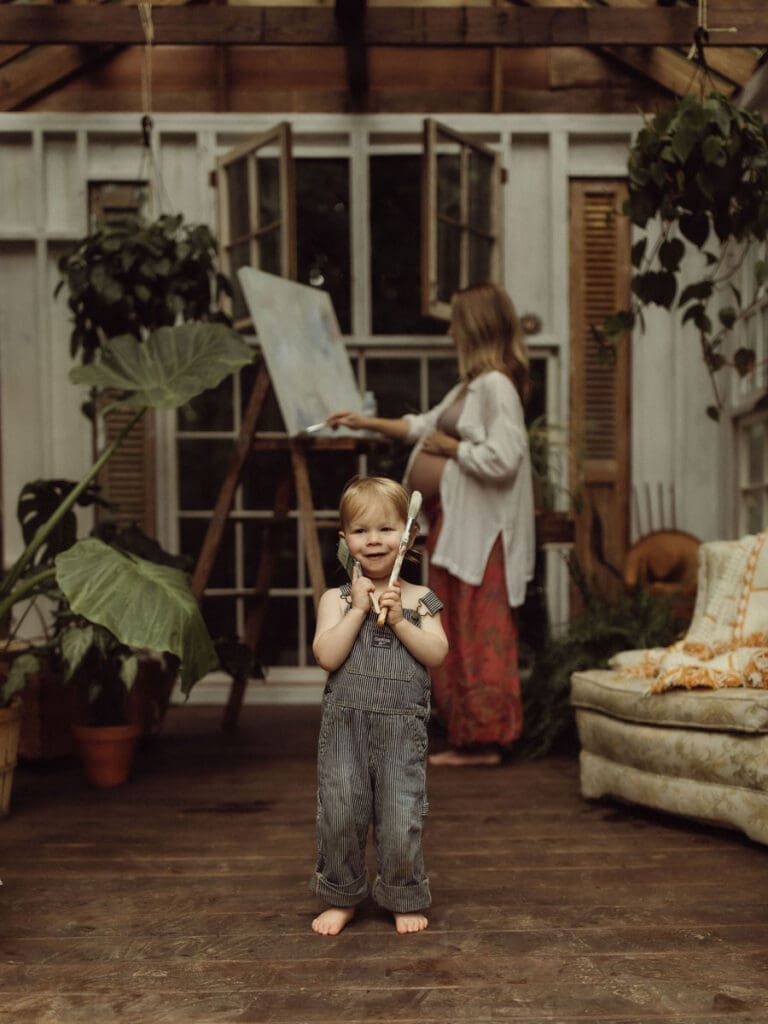 A toddler boy holding paint brushes to his face while his pregnant mom is painting on an easel behind him.  
