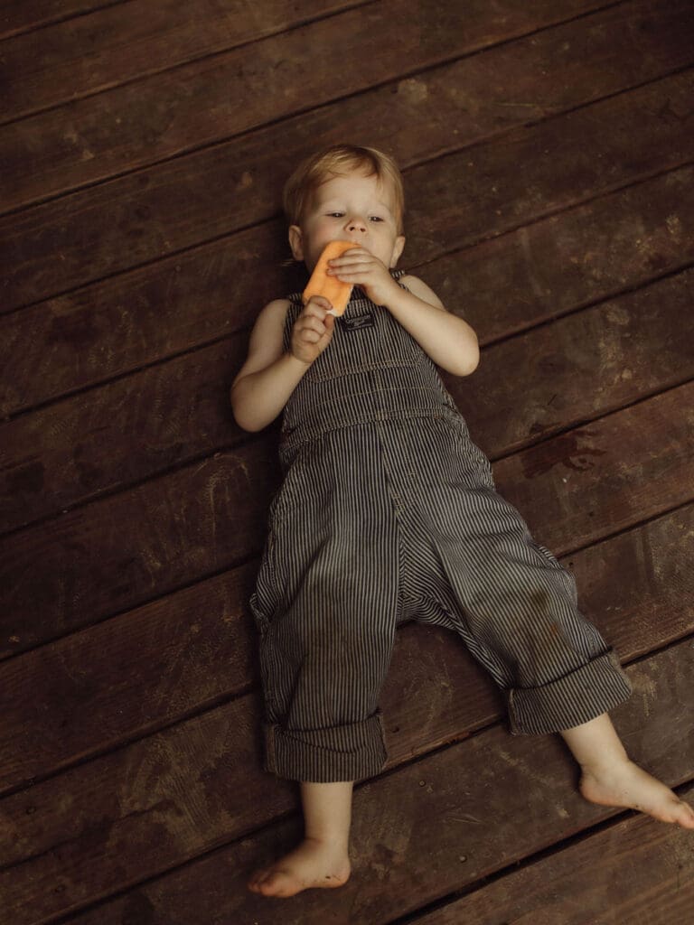 A little boy in dirty overalls laying on the wood flooring of Das Hinterhaus eating an orange popsicle. 