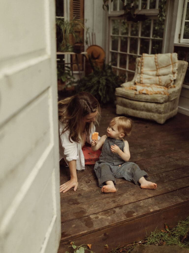 A toddler offering a bite of his orange popsicle to his mother while sitting on the floor at Das Hinterhaus of Marietta, GA. 
