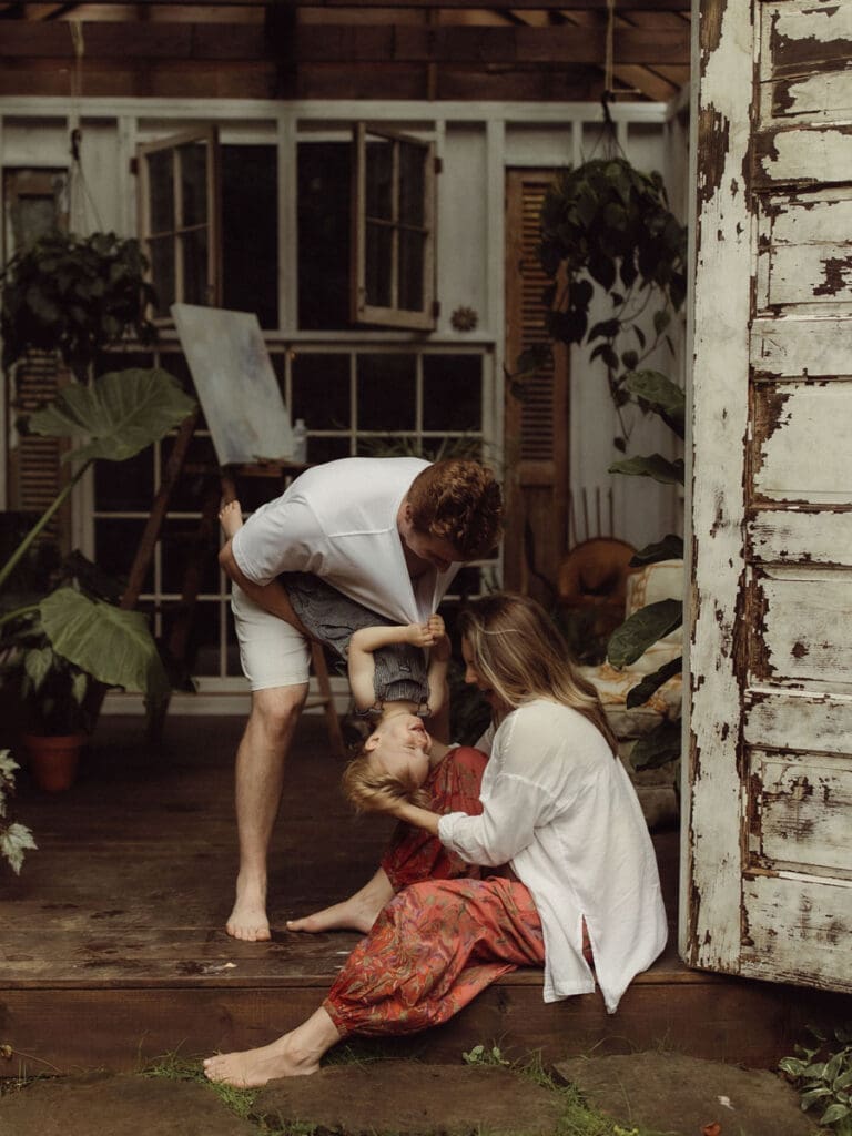 A dad tipping his toddler upside down so his mom can give him a kiss while she sits on the ground. 
