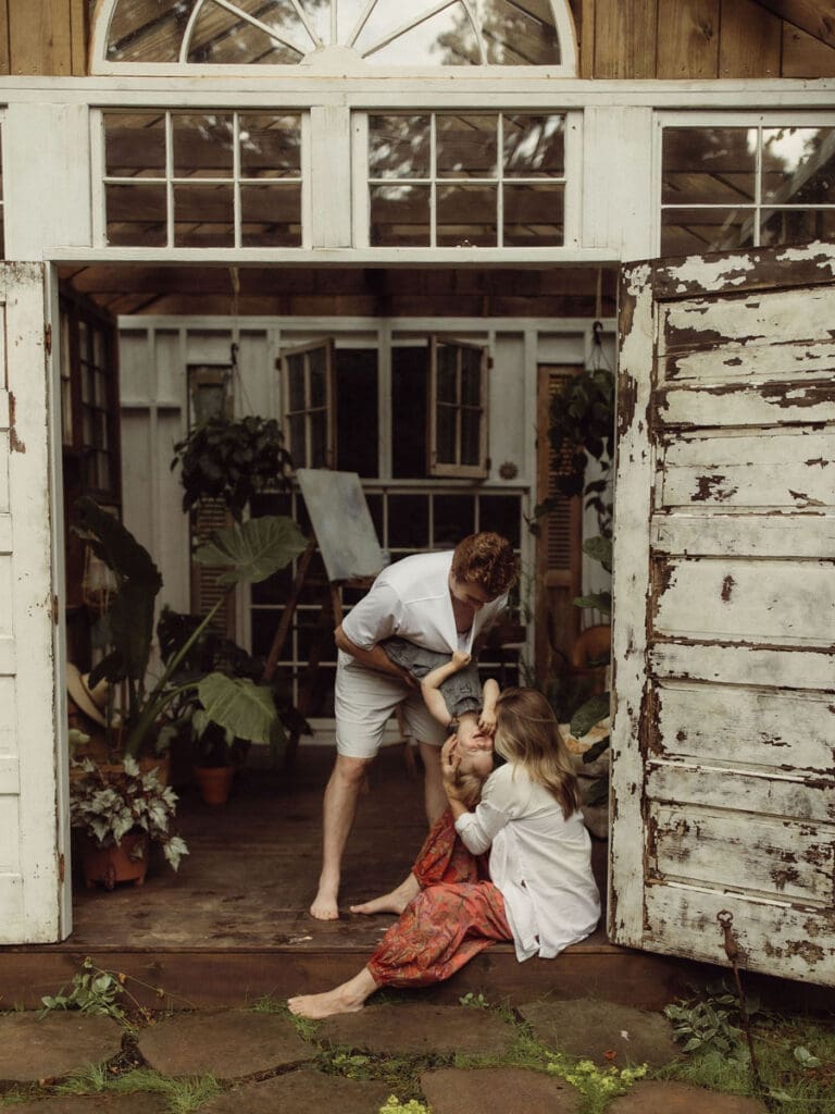 A dad holding his toddler upside down so his pregnant mom can give him a kiss while she sits on the ground. 