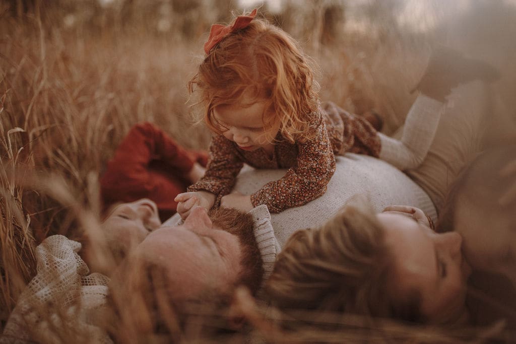 A toddler girl with curly red hair laying on her father and playing with his beard. 