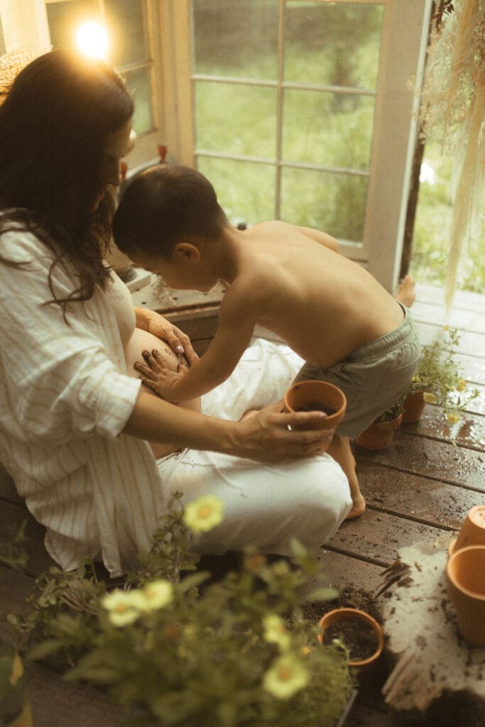 shot in a vintage greenhouse made of old windows, expectant mother and her son pot flowers. Little boy leaves muddy handprints on mom's pregnant belly.