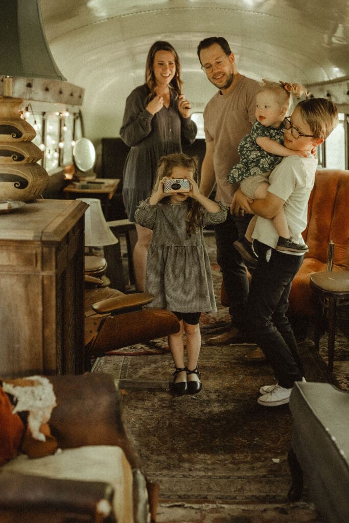 parents and three young kids dance in a renovated old school bus. five year old little girl holds disposable camera and shoots back at the photographer