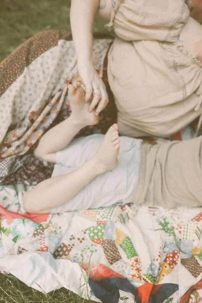 detail shot of a mom tickling the foot of her young son. They're both laying on a quilted, colorful blanket on top of tall green grass. Mom wears a hand maid long, patchwork skirt and little boy on his stomach with his feet kicked up so mom can tickle the soles of his feet