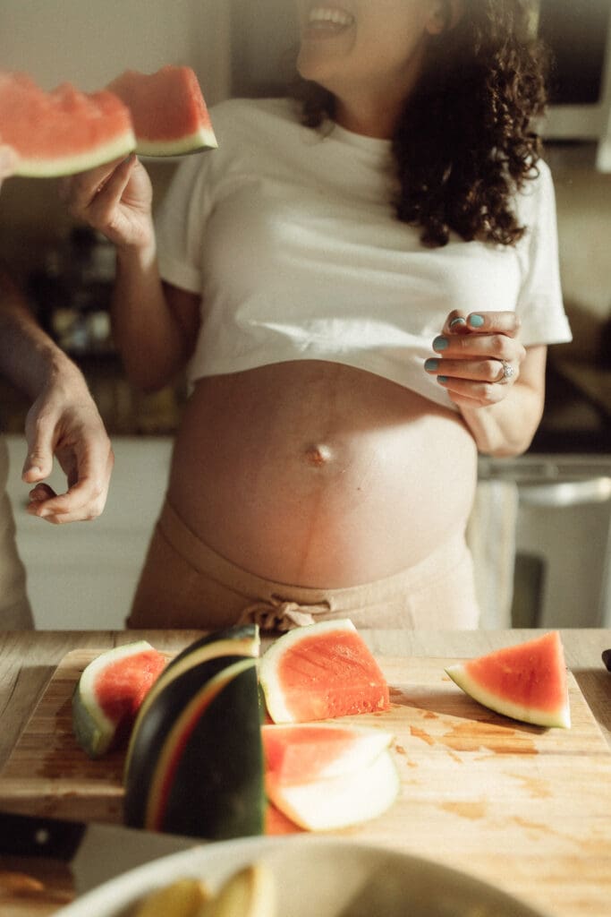 expectant mom and dad eat watermelon. Mom's exposed belly is covered in drips from the juicy watermelon and she smiles with a bug toothy grin up at her husband.