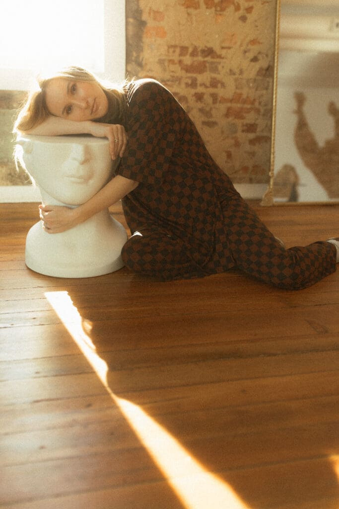 woman wearing a matching brown and black checkered outfit rests her head on a table shaped like a head. a bright stream of sunlight pours in directly behind her from a window. she rests one hand under her head and wraps the other around the neck of the head table.