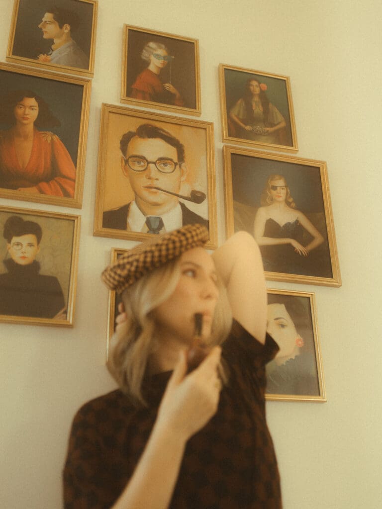 woman with a checkered shirt and hat on sitting in front a gallery wall of photos. The biggest shot in the gallery wall is a man with a pipe in his mouth, so the woman being photographed also has a pipe hanging from her mouth. Image is intentionally backshot with the focus falling on the portrait of the man with pipe rather than the subject in the foreground