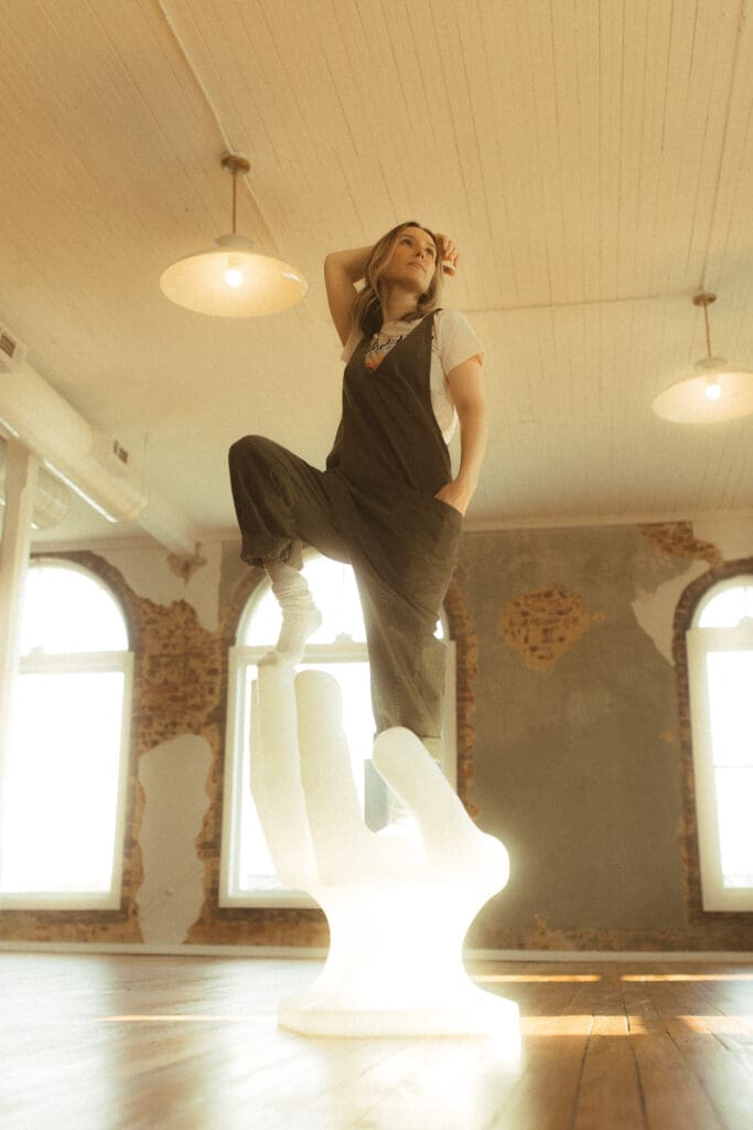woman stands on a hand-shaped chair. One foot on the palm with her other foot propped up on the middle finger of the chair. The sun shines through the white chair making it look like it's glowing. Her hand is thrown back behind her head and her chin is tilted up, photographer shooting up at her making her look long and strong.