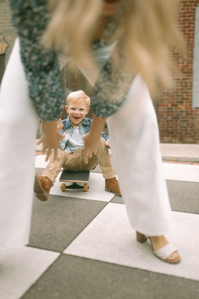 little boy sits on skateboard and mom reaches down between her legs to pull him through her wide-stretched stance. Image is shot through moms legs. you see the little boy with bright blonde hair, glasses and missing front teetn smiling and reaching for his mom's hand. You see mom's blond hair falling and framing the top of the shot.