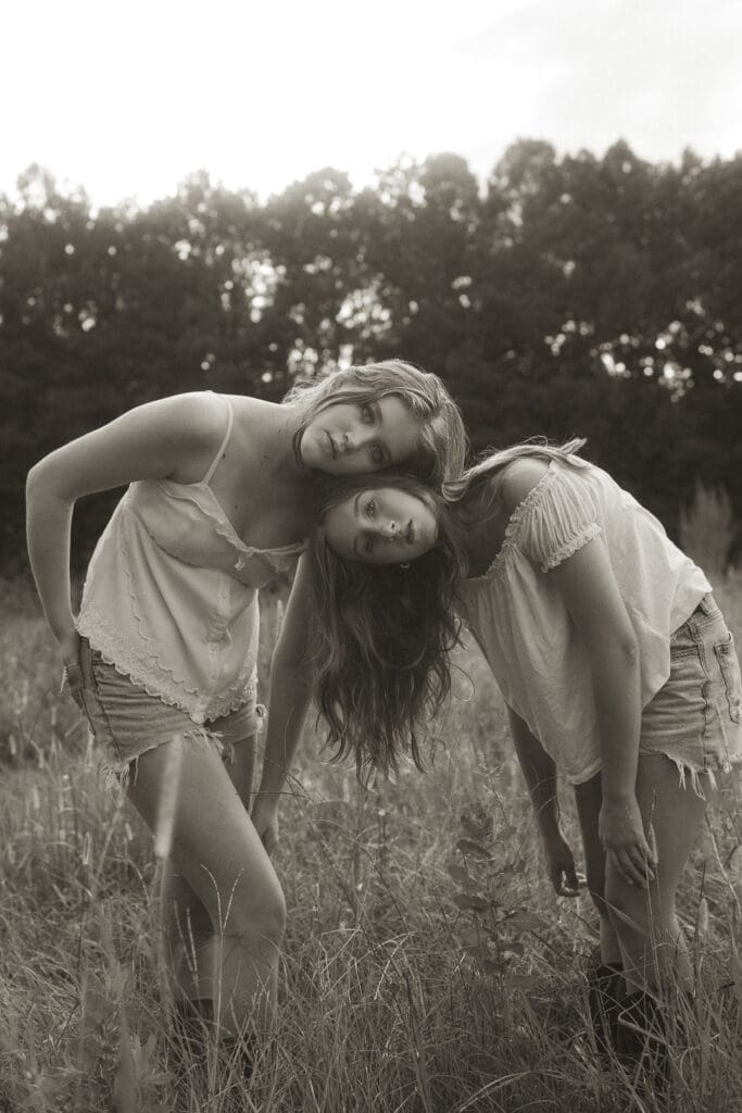 two teenage girls in black and white. shot in a field of tall grass, girls wearing cut off jeans shorts and bent over with their heads resting on each other and arms dangling. a broken, slouchy pose but they have serious gazes on their faces, piercing straight into the camera