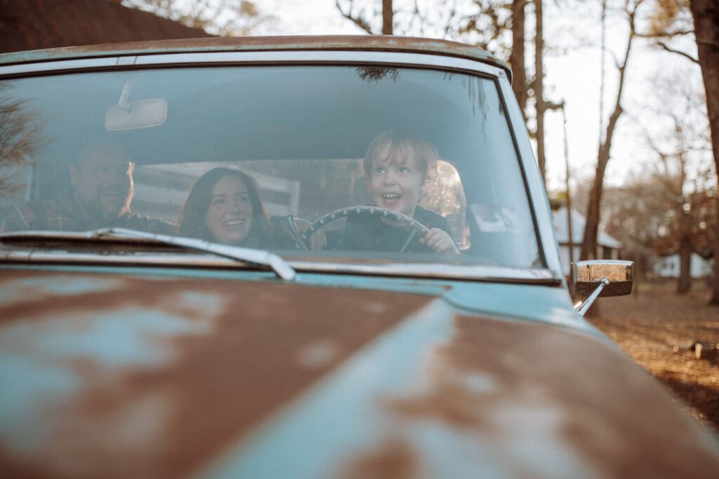 parents and their young boy in the front cab of an old vintage ford truck. Little boy smiles as he pretends to drive and parents watch and smile. shot with the camera resting on the hood of the truck, the hood acts as a leading line up to the smiling boy at the wheel