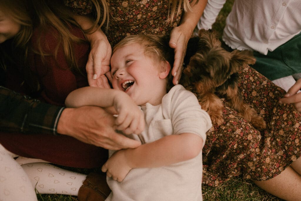 family sits on. the grass with the youngest child's head in everyone's lap. mom's hands frame is smiling cheeks and dad's hand reaches over to tickle the boy. family yorkie sits next to the boy's head, in mom's lap and watches intently