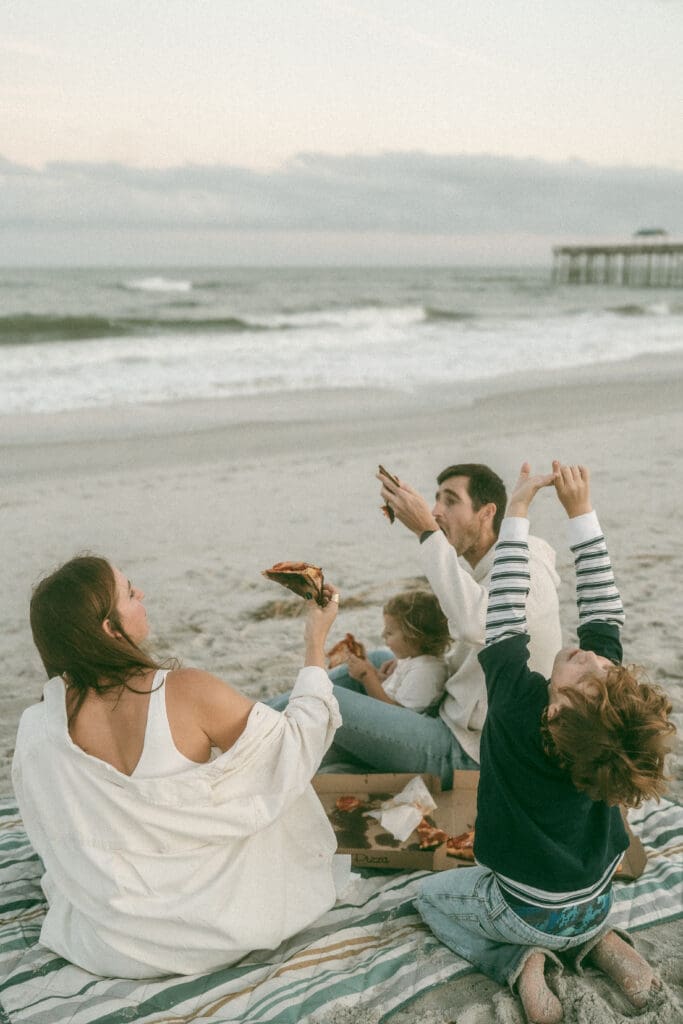 family sits on a beach in jacksonville, fl and eats pizza. They're holding the pizza in the air in a celebratory way.