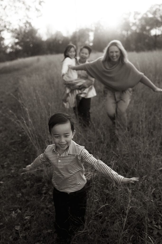 little boy flies like an airplane with arms stretched wide through a field of tall grass, as his moms follow suit behind him- one mom also with outstretched arm, the other holding his slightly older sister. Shot in black and white, focused on the boy in the foreground with focus falling off towards the smiling moms