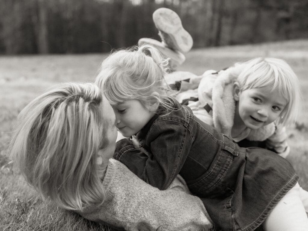 mom lays on her back in the grass with one young daughter laying on top of her, snuggling her nose to nose. Sister comes from the other direction and lays across moms legs and peeks at the snuggles between big sister and mom. black and white image