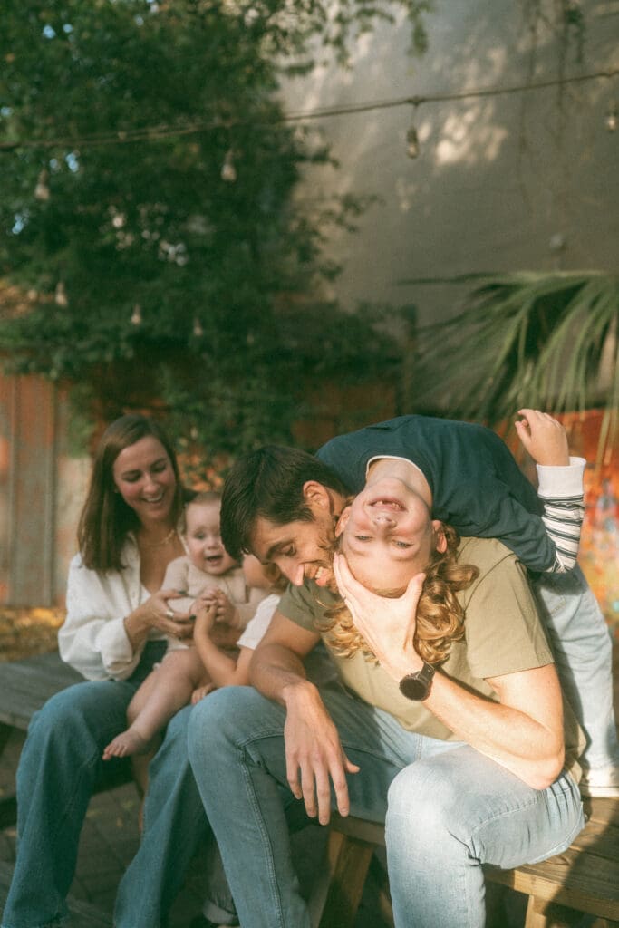 young family sits on a picnic table. Mom loves on baby sister while big brother hangs his head backwards over dad's shoulder- face towards the camera with a big, toothless grin. color image with a direct sliver of sunlight right over the dangling boy's head. 
