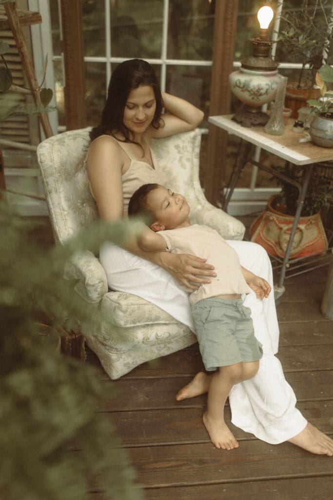 mother sits in a vintage chair inside a greenhouse made of old windows and snuggles her young son as he leans back against her legs.