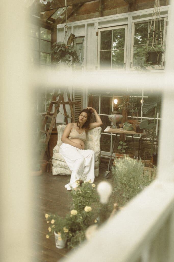 portrait of a pregnant mother sitting in a vintage arm chair inside a greenhouse built from old windows. Shot from the outside, looking in.