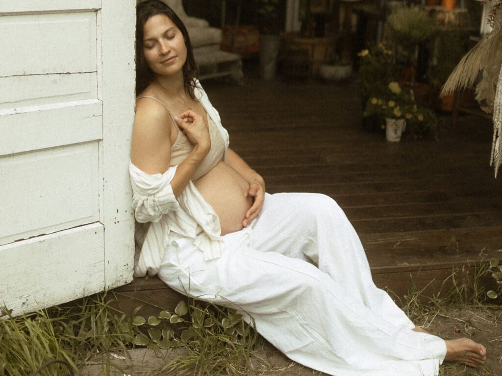 expectant mother sits in the opening of a greenhouse, leaning against the door frame. Her pregnant belly is exposed, she closes her eyes with a sweet, subtle smile on her face, rests one hand on her belly and the other over her heart and stretches her out her legs.