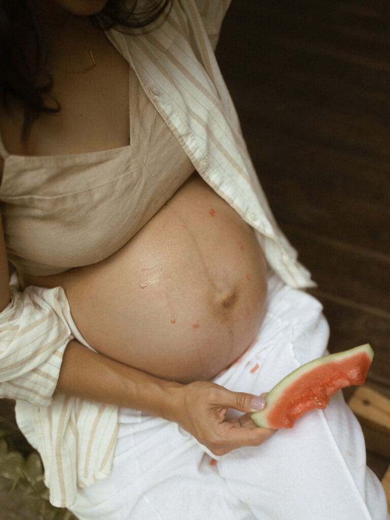 close up shot of a shirtless pregnant belly covered in drips of watermelon juice. Mom holds a piece of almost eaten watermelon wedge in her hand.