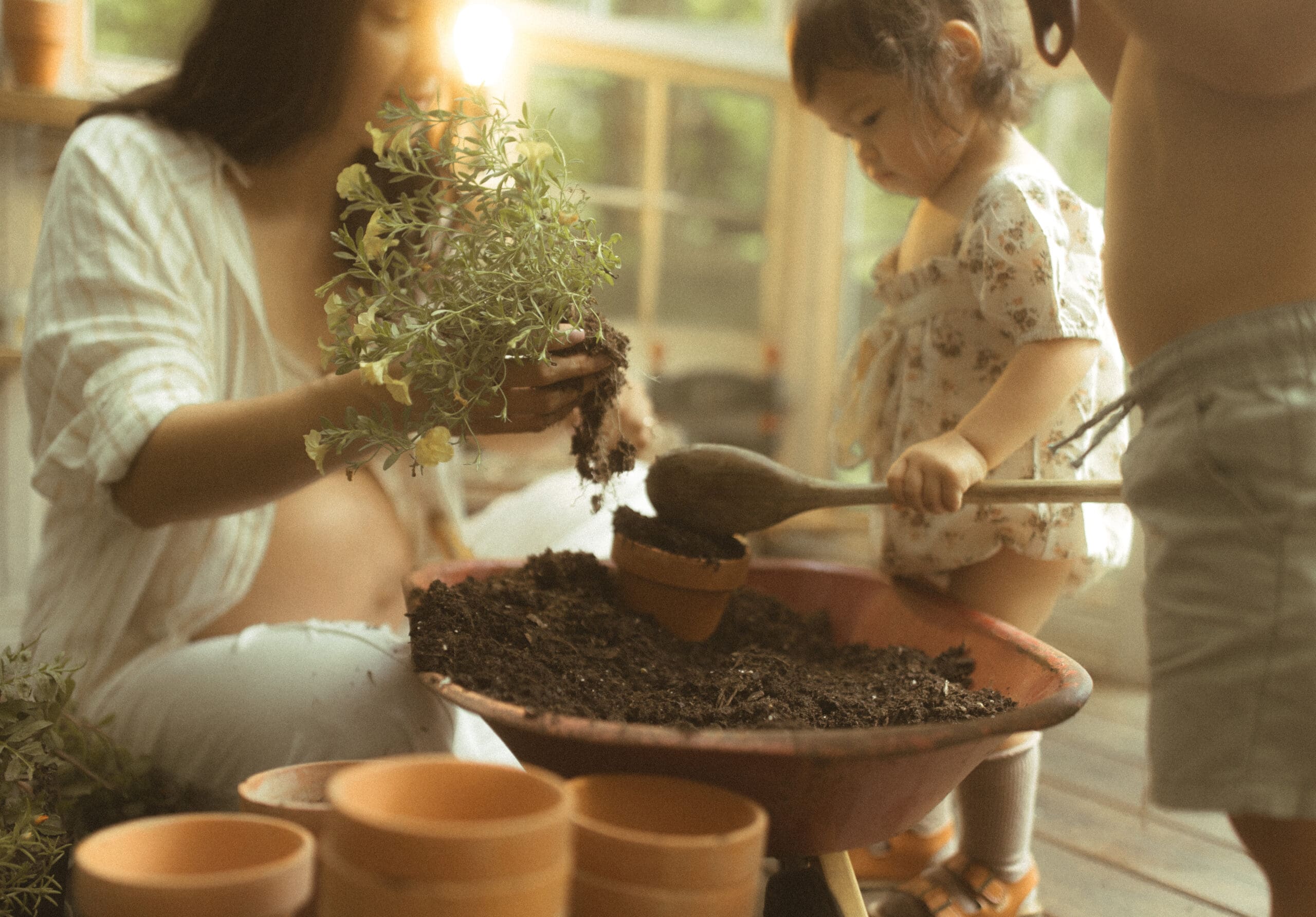 a pregnant mother and her two young children using potting soil and small red, clay pots to pot flowers inside a vintage greenhouse.
