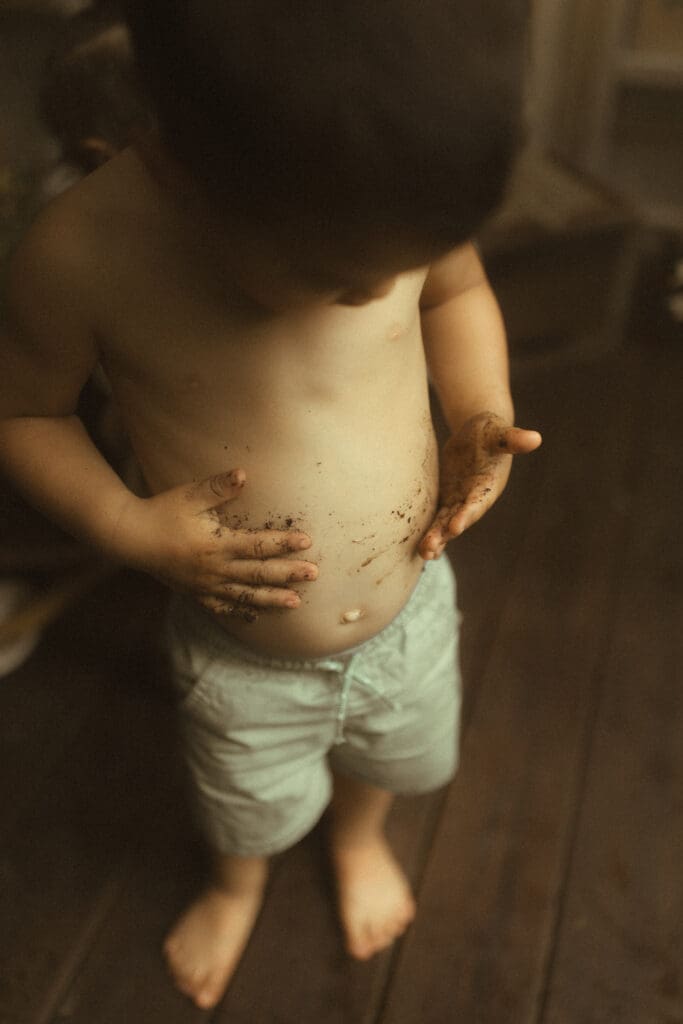 shirtless little boy takes his hands that are covered in potting soil and makes handprints on his bare belly.