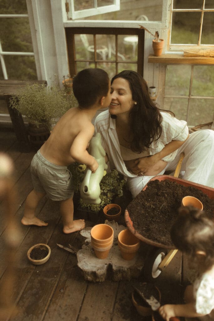 mom, son and daughter digging in a small wheelbarrow of potting soil and potting flowers into small clay pots. Shot inside a greenhouse built of old windows.