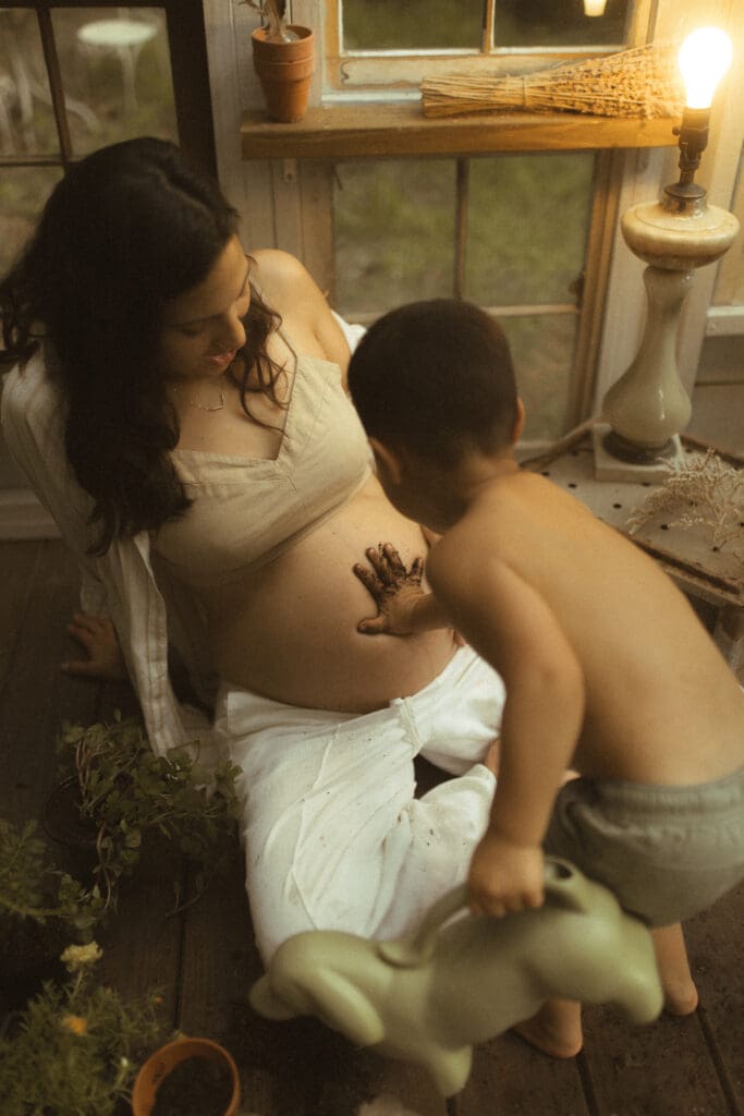 little boy uses his potting soil covered hands to make a handprint on the exposed belly of his very pregnant mother.