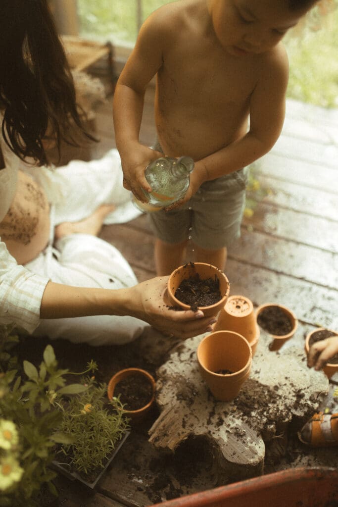 mom, son and daughter digging in a small wheelbarrow of potting soil and potting flowers into small clay pots. Shot inside a greenhouse built of old windows.