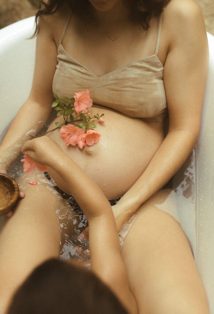 detail shot of a bare, pregnant belly with some pink azalea flowers resting on her belly. Mom sits outside in a cast iron tub filled with water. The head of her son can be seen adding additional azalea petals to her stomach.