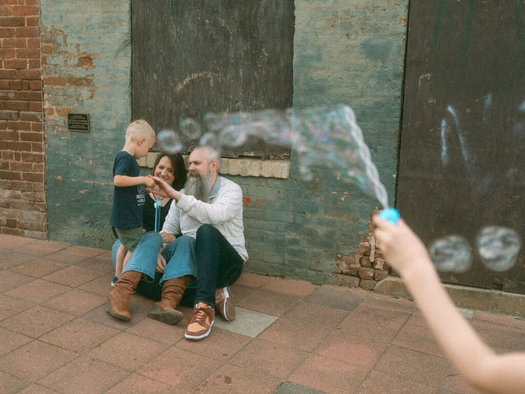 mom and dad help the youngest blow bubbles while big sister frames them in the foreground with a wand of bubbles