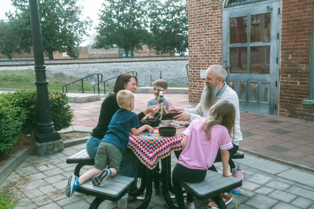 whole family eats ramen and sushi around a picnic table in an alley in downtown acworth. Middle brother uses the camcorder to document