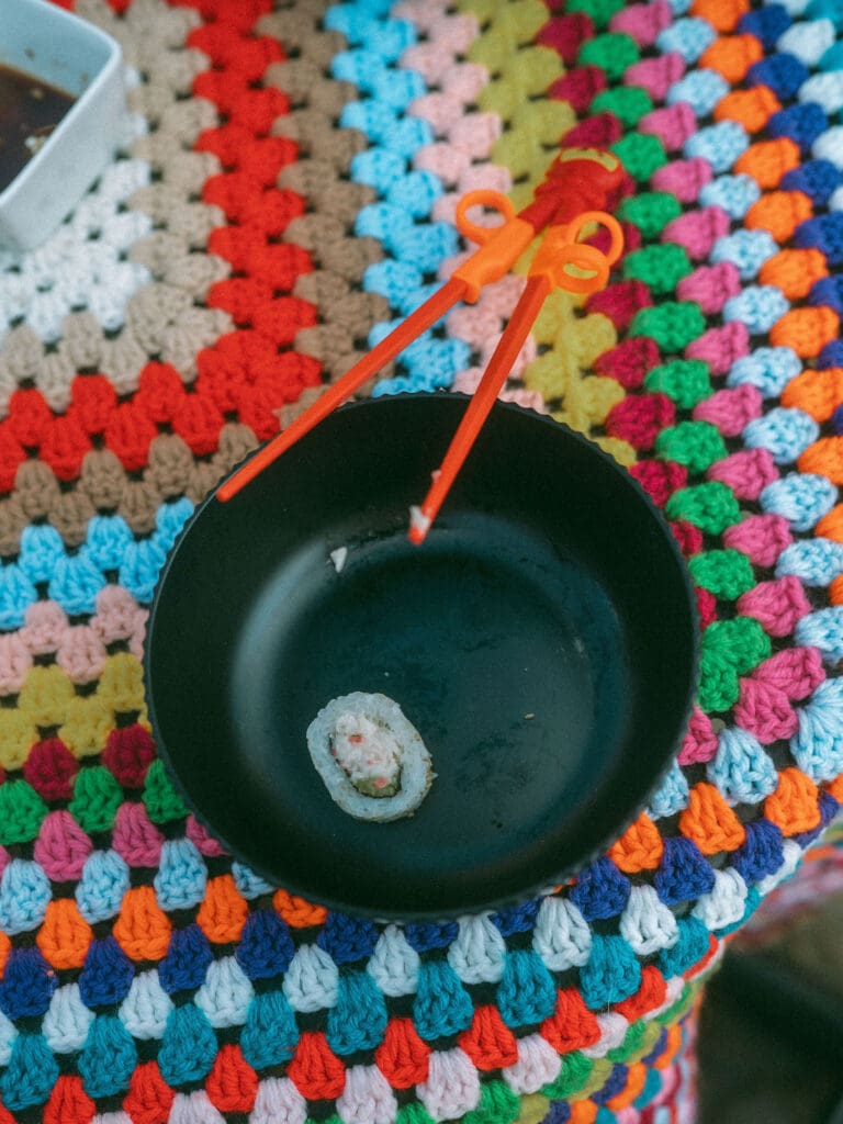 one last piece of sushi sits in a black bowl on a colorful tablecloth with a red pair of children's chopsticks