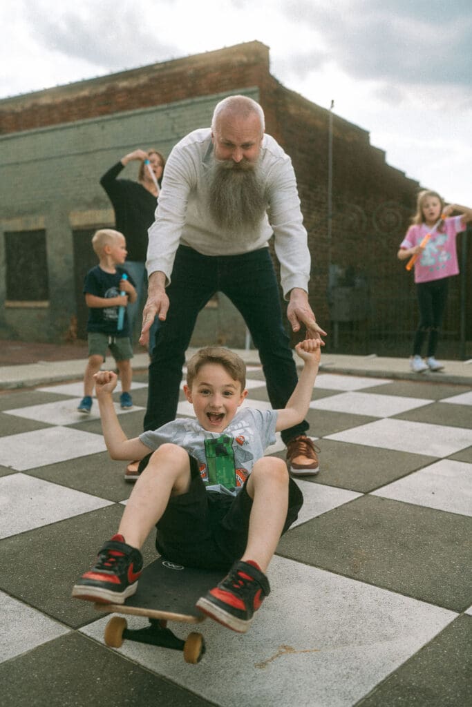middle son sits down on skateboard and is pulled through dads legs as the rest of the family plays in the background