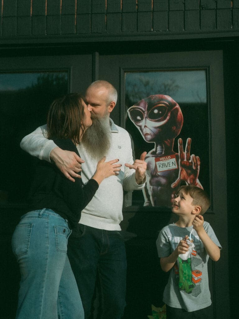 mom and dad kiss in front of a door with a cardboard cutout of an alien- little boy eats ice cream next to them and stares up at their kiss
