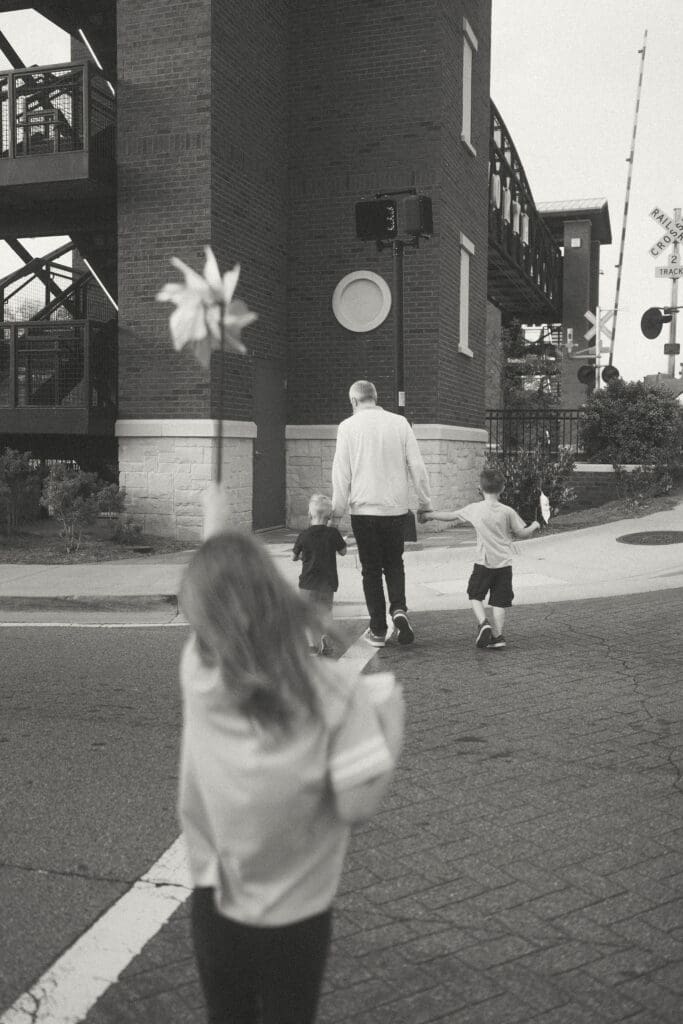 dad holds hands of boys while they cross the street and sister stays behind with her pinwheel held high in the air